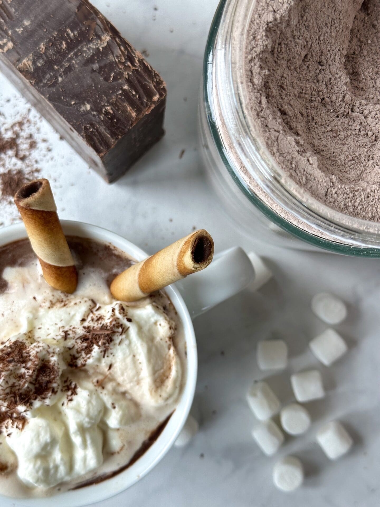 IMG_3623 A mug of hot chocolate is seen from above, topped with whipped cream, chocolate shavings and cookie rolls. A jar of the hot chocolate mix, a pile of mini marshmallows and a large block of dark chocolate are seen in the background