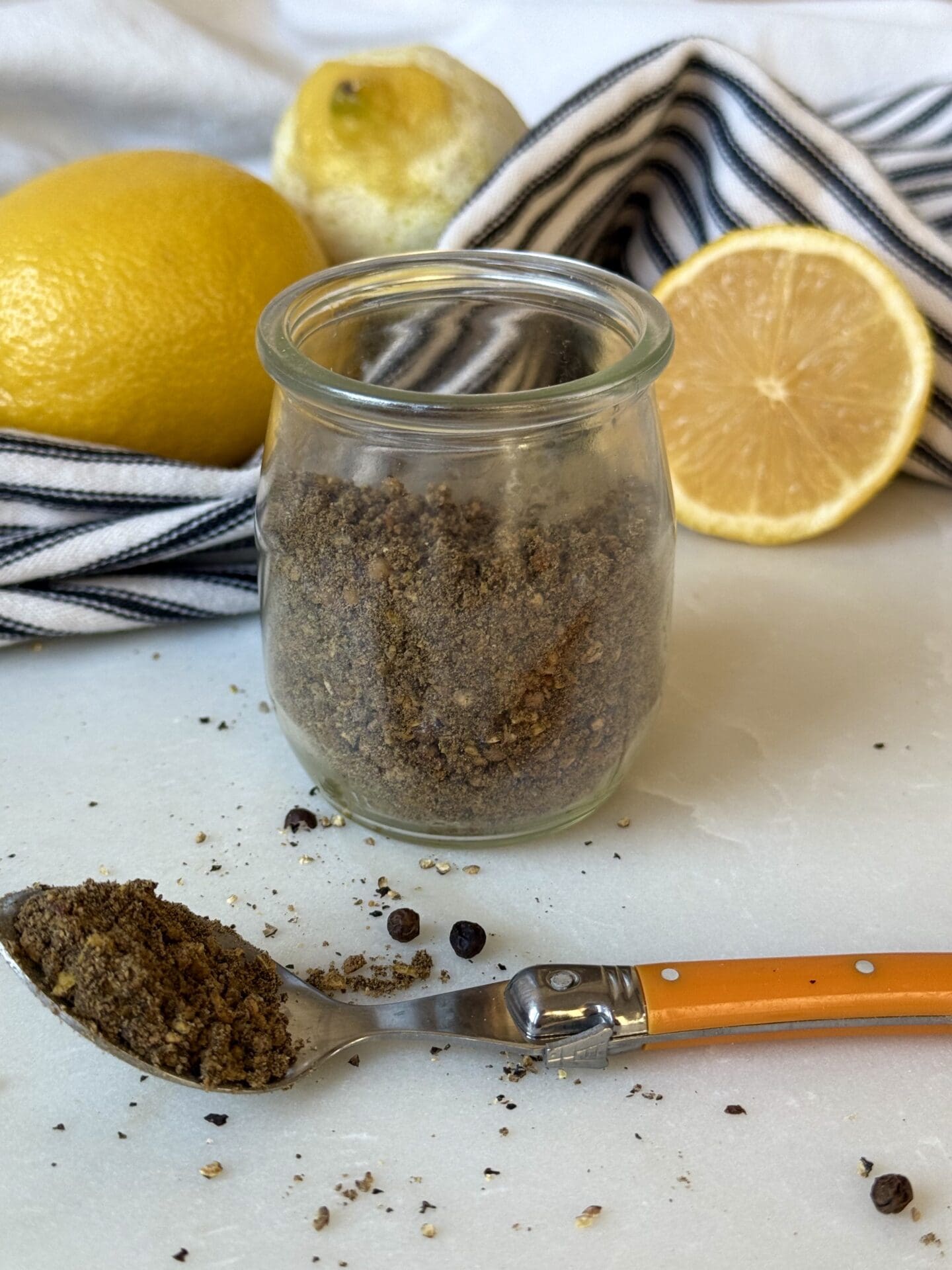 IMG_4129 A jar of homemade lemon pepper seasoning sits on a white marble table. A spoonful of the spice blend spills out onto the table in the foreground. Fresh lemons and a black and white striped cloth are seen in the background.