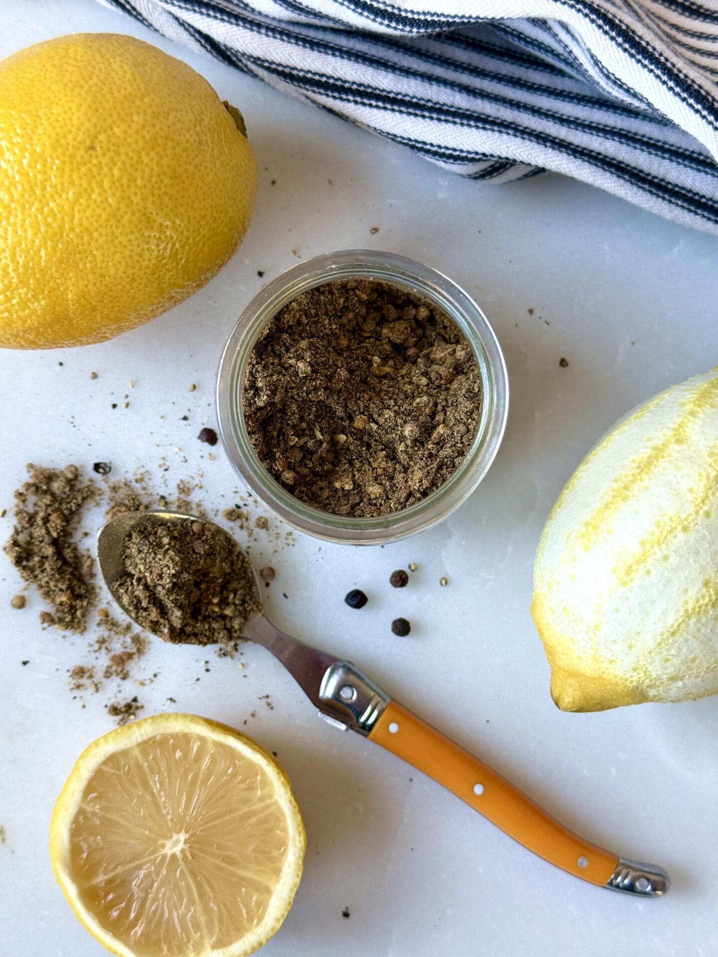 IMG_4136 A jar of Homemade Lemon Pepper Seasoning is seen from above, with a spoonfull of the spice mix spilling onto the white marble table. Fresh lemons and peppercorns surround the image.