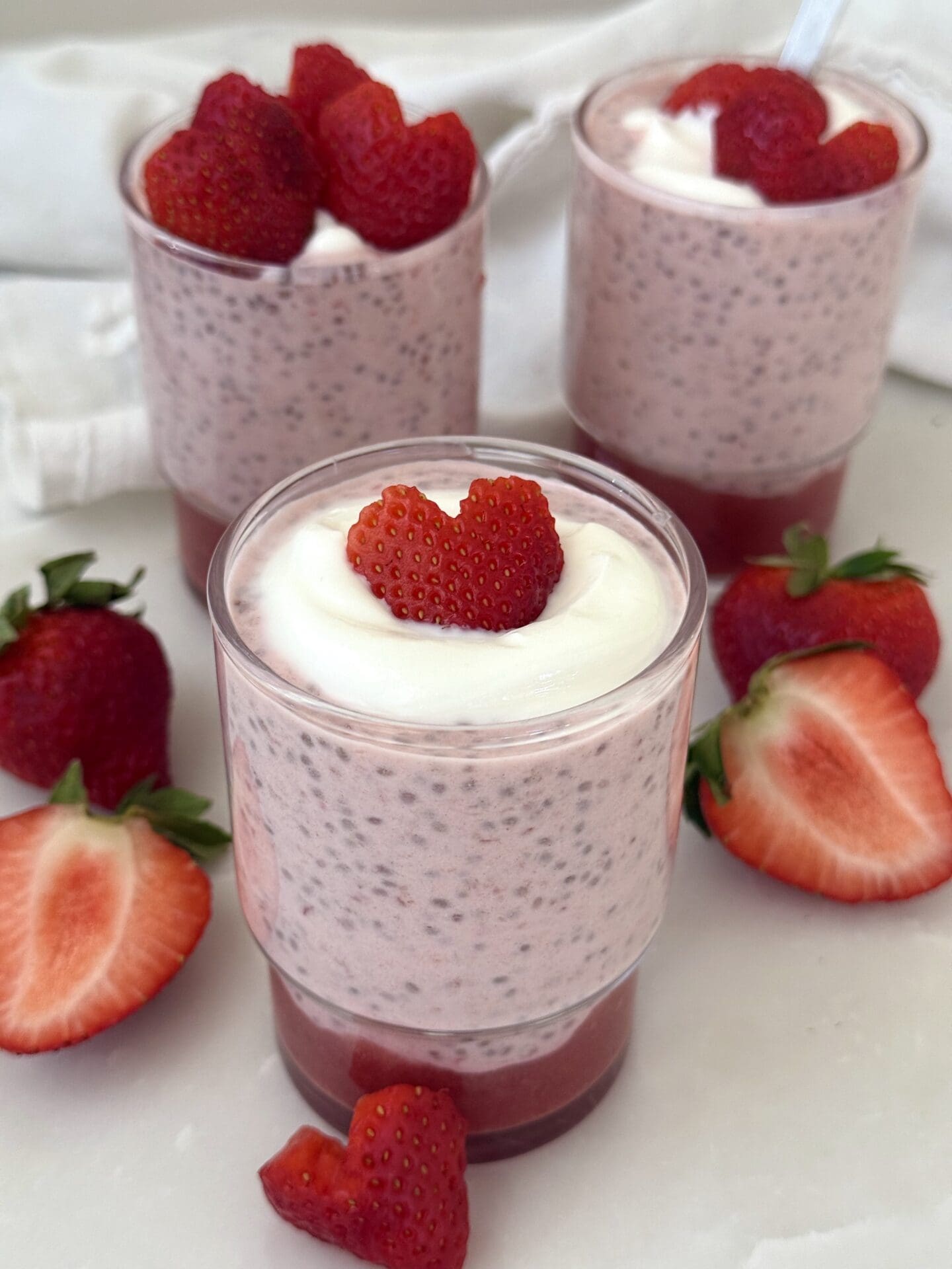 IMG_4187 A trio of glass jars hold individual servings of strawberry chia seed pudding, sitting on a white marble table surrounded by fresh strawberries