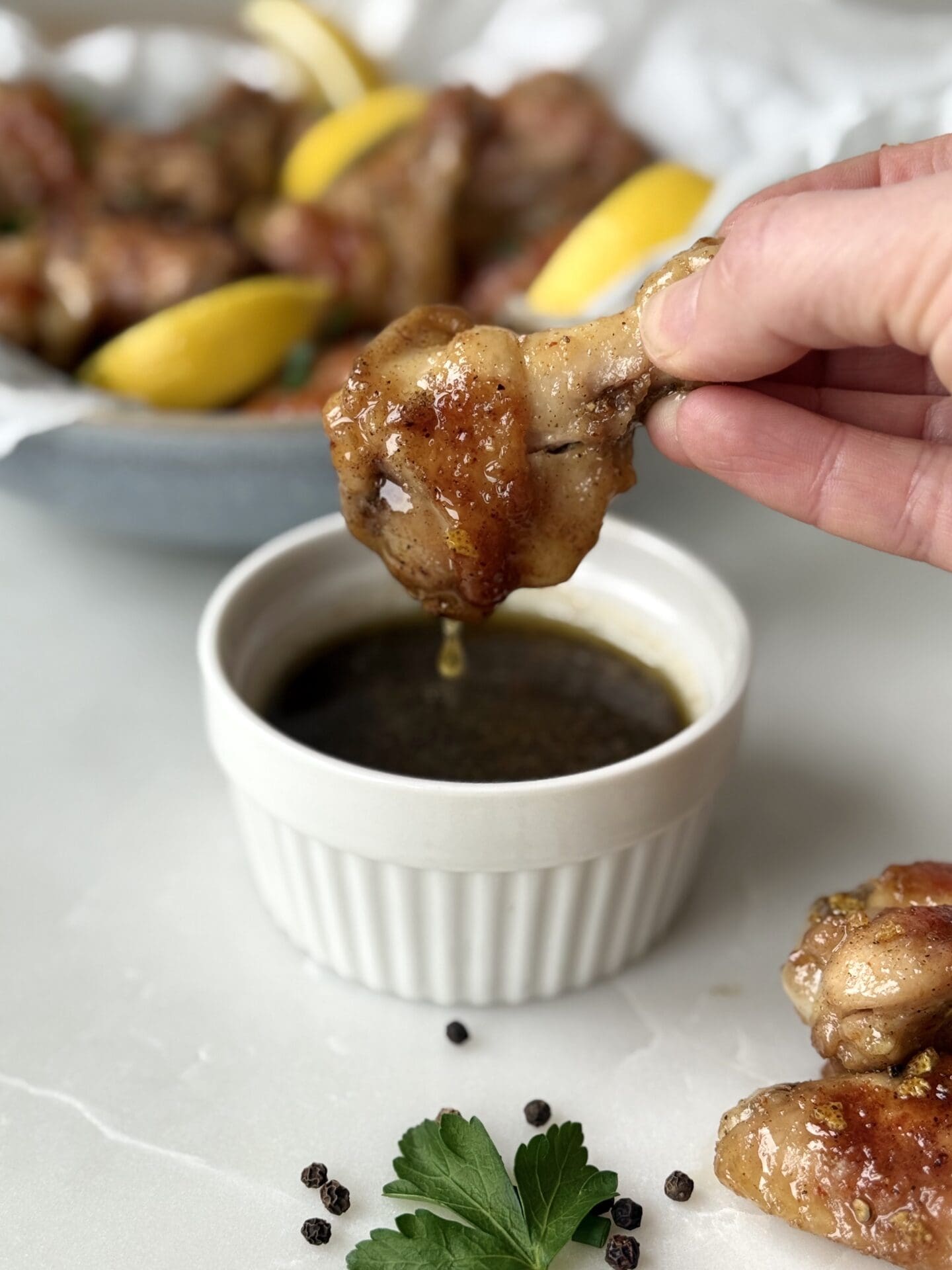 IMG_4273 A hand is seen dipping a lemon pepper chicken wing into a bowl of sauce. A platter of wings and lemon wedges is seen in the background.