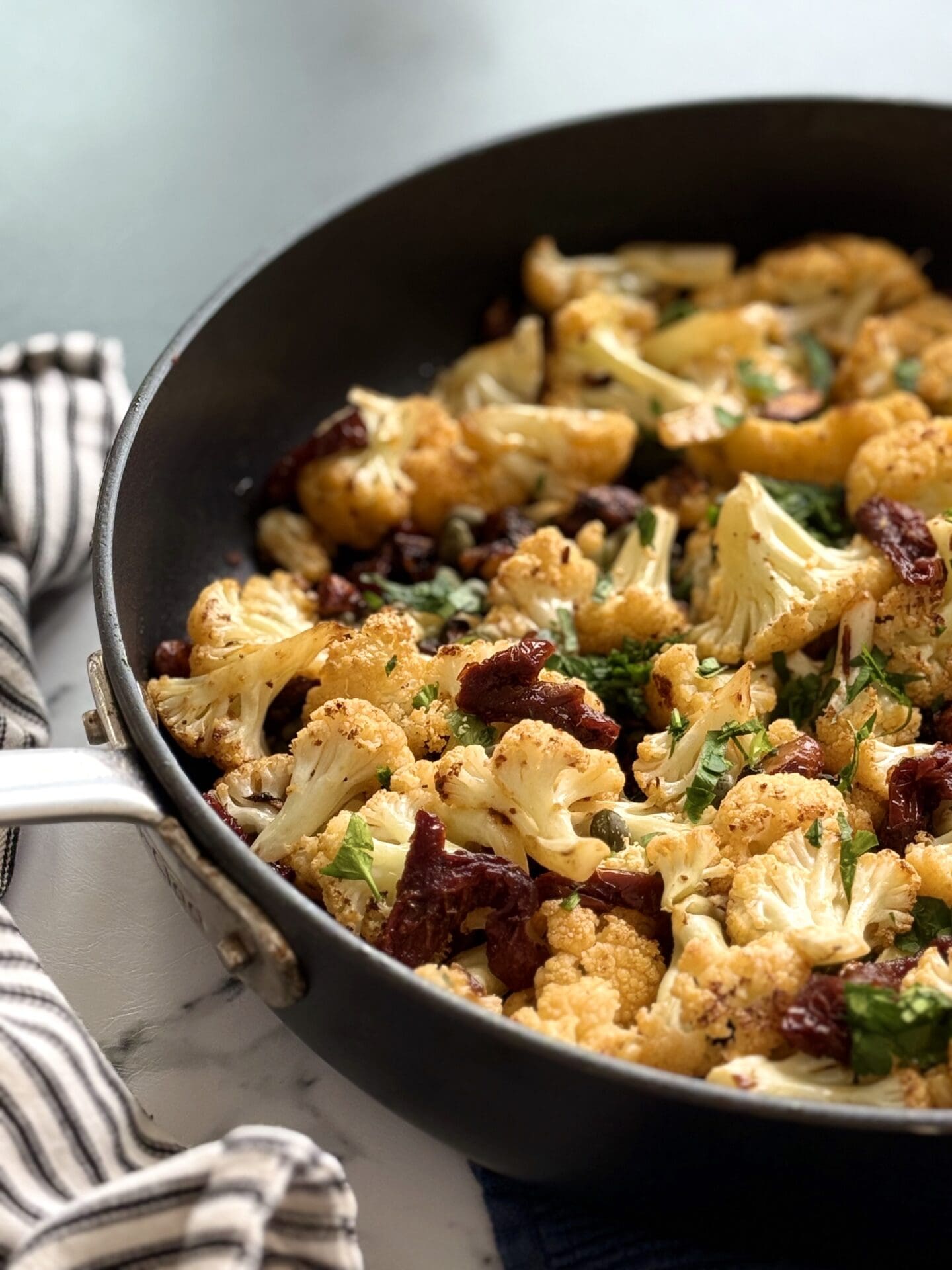 IMG_4434 A large black skillet full of Roasted Cauliflower with Sundried Tomatoes, Capers and Crunchy Almonds is seen up close with a black and white striped towel wrapped around the skillet handle.