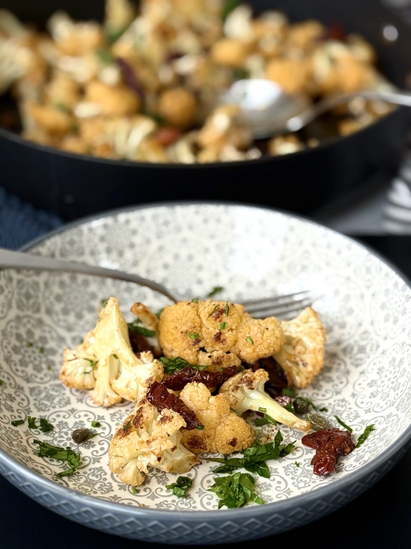 IMG_4441 A serving of Roasted Cauliflower with Sundried Tomatoes, Capers and Crunchy Almonds is seen in the foreground in a grey patterned bowl. In the background a black skillet full of this cauliflower dish can be seen.