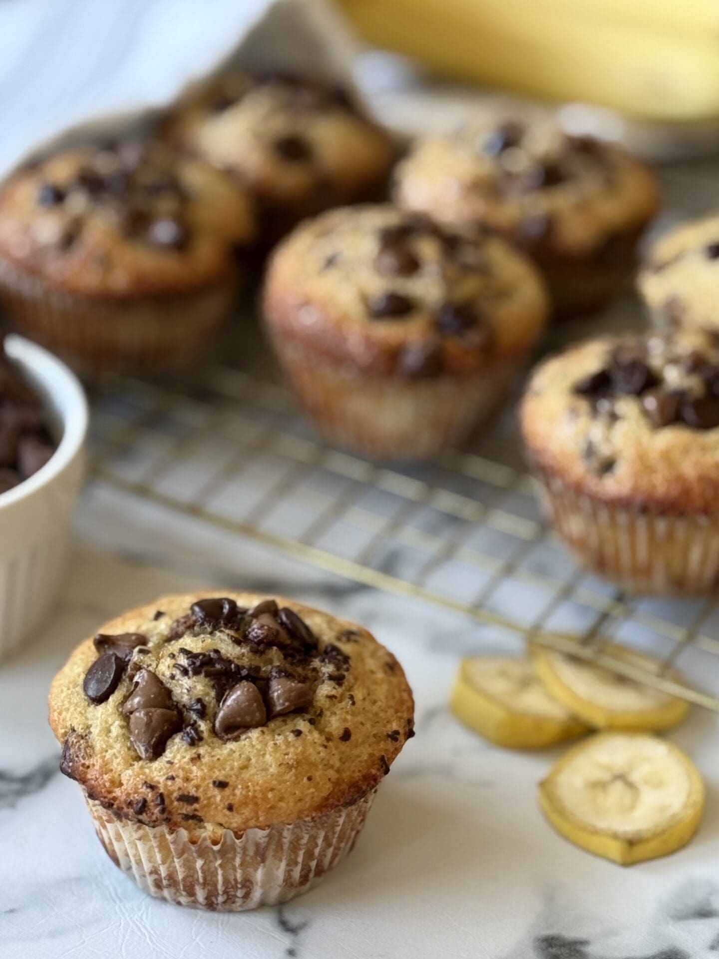 IMG_4511 A Sour Cream Banana Chocolate Chip Muffin is seen in the foreground, with other muffins, banana slices, whole bananas and chocolate chips seen in the background.