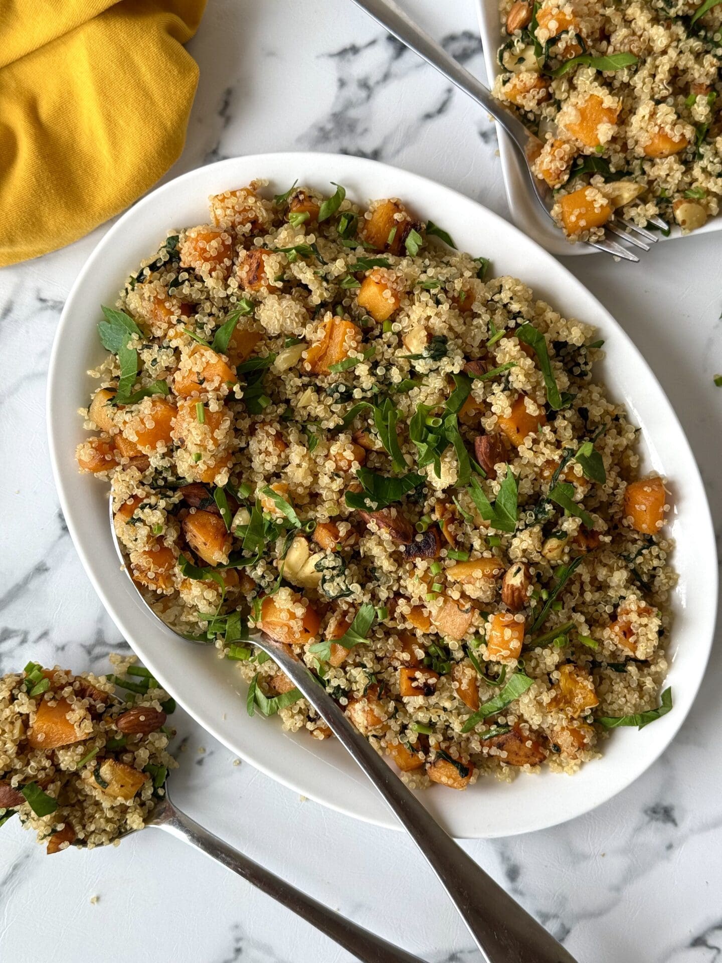 IMG_4705 A platter of quinoa salad with butternut squash, spinach and crunchy almonds is seen from above on a white marble counter, surrounded by serving utensils and a yellow dinner napkin.