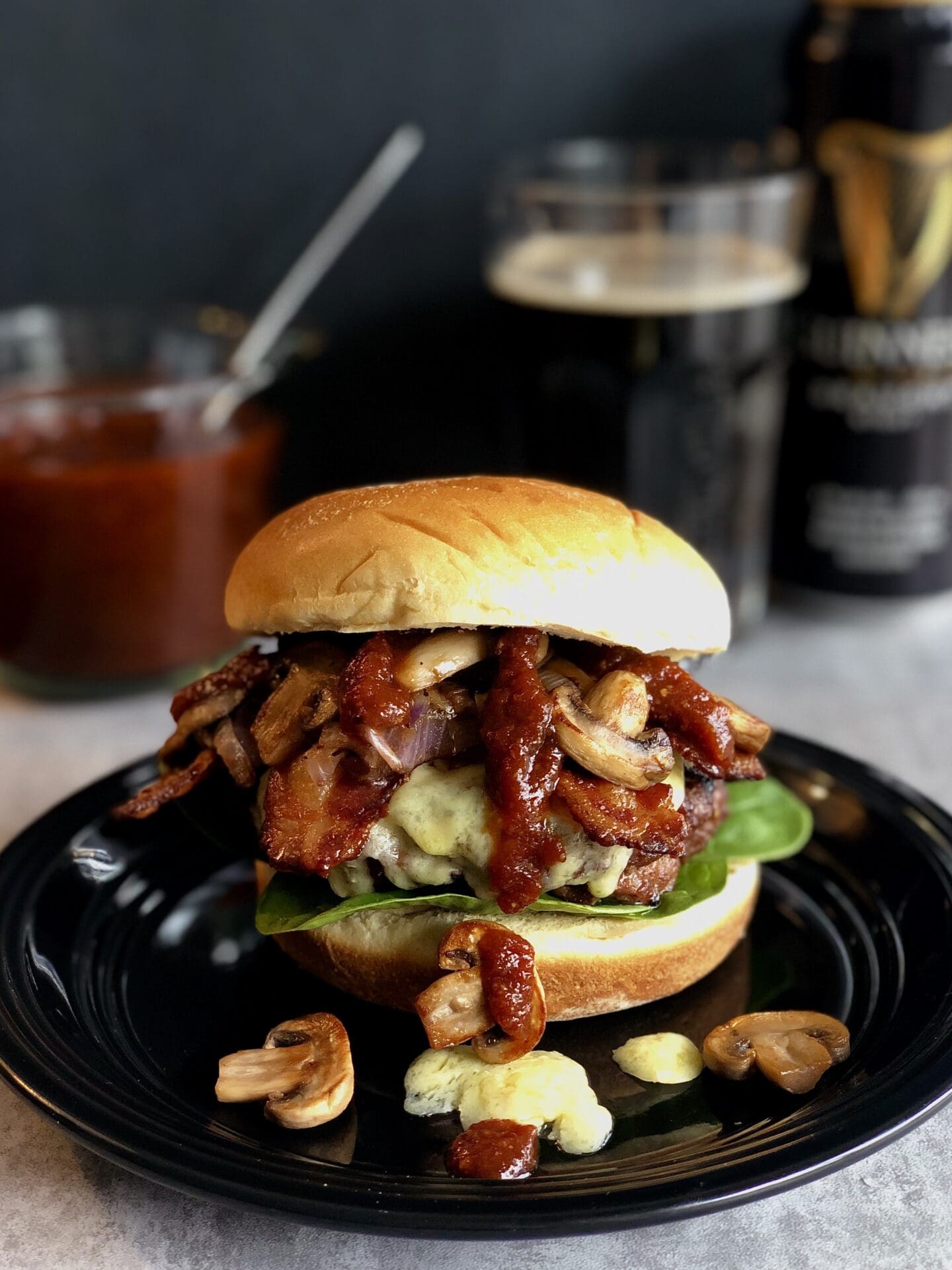 IMG_4171 A Guinness Bacon Burger with Mushrooms and Irish Cheddar is seen on a black plate. Guinness Barbecue sauce and a can of Guinness beer can be seen in the background