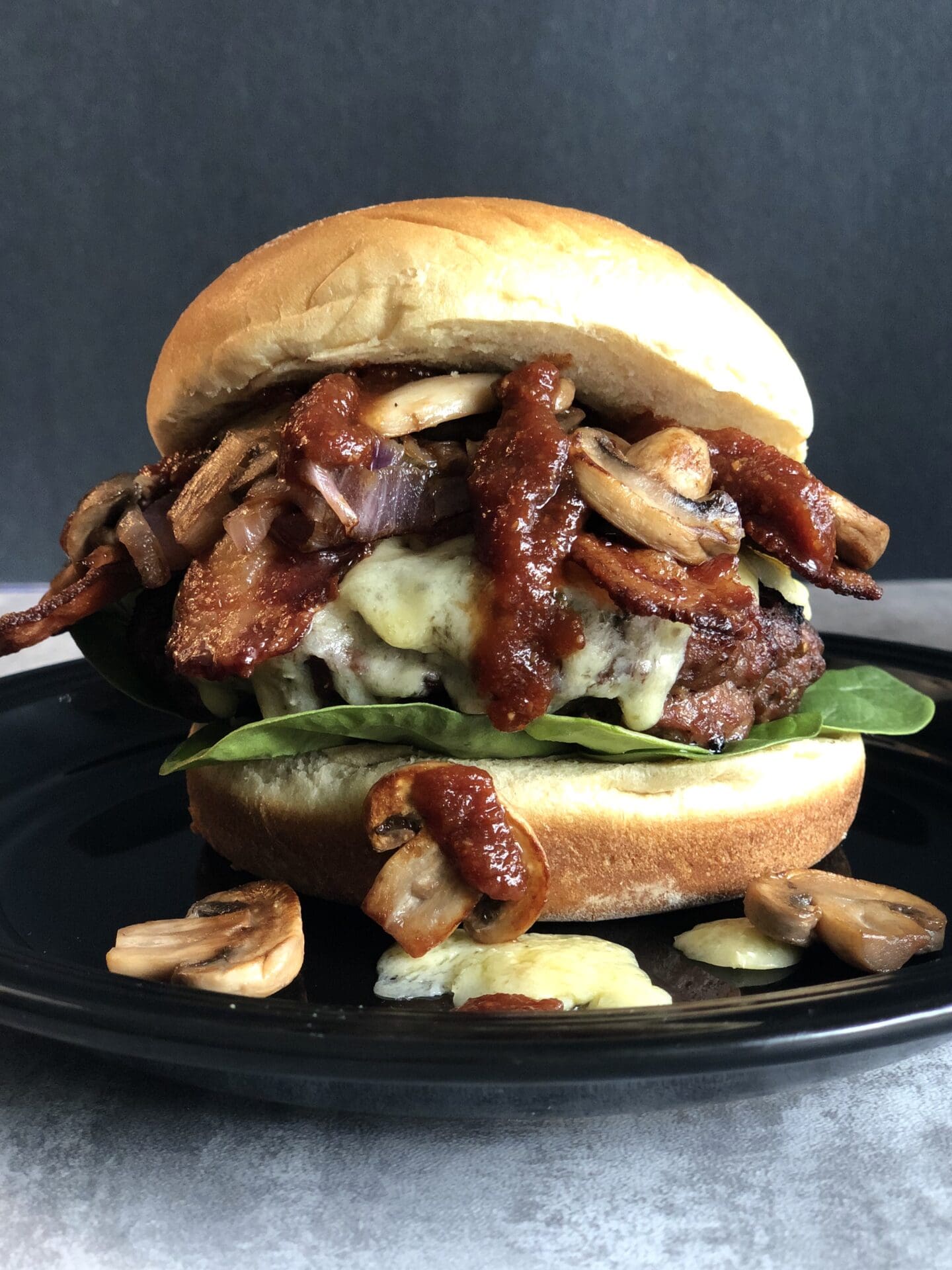 IMG_4174 A Guinness Bacon Burger with Irish Cheddar and Mushrooms is seen close up. Guinness barbecue sauce drips down and golden sautéed mushrooms tumble out of the burger onto the black plate underneath.