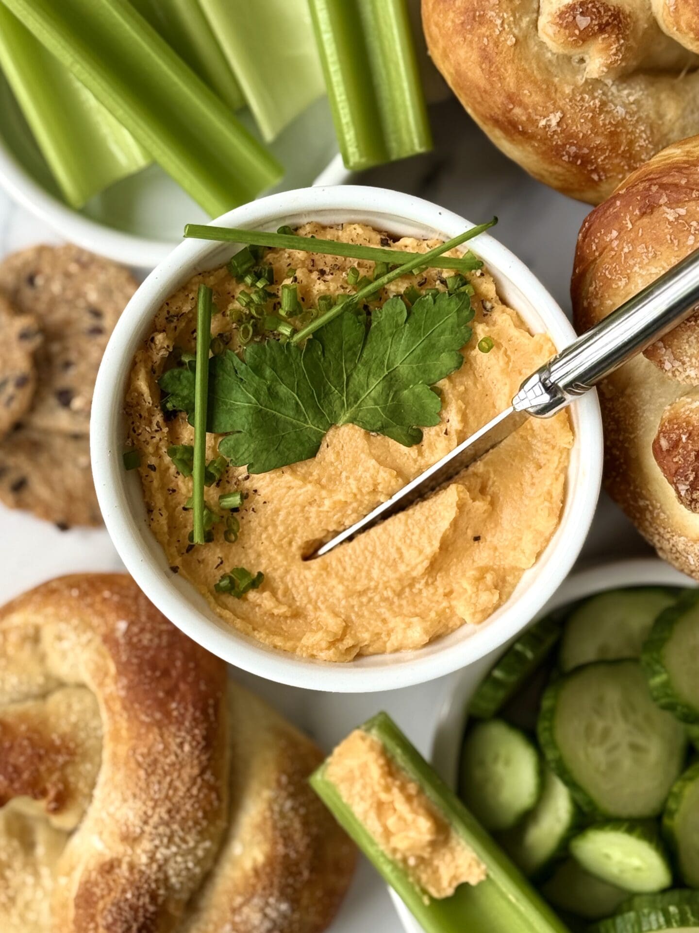 IMG_4637 A dish of Pub Style Beer Cheese is seen from above, garnished with fresh herbs and surrounded by green vegetables and soft pretzels.