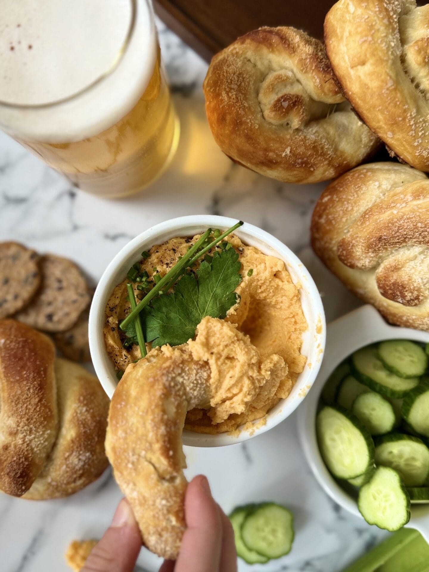 IMG_4653 A bowl of Creamy Pub Style Beer Cheese is seen from above surrounded by a cold beer, cucumber slices and crackers. A hand is seen reaching in and dipping a soft pretzel in the Beer Cheese.