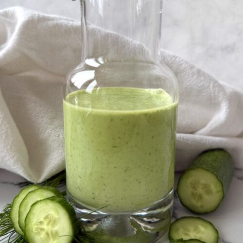 A glass bottle of Creamy Cucumber Dill Salad Dressing is seen on a white background, surrounded by fresh dill and sliced cucumbers
