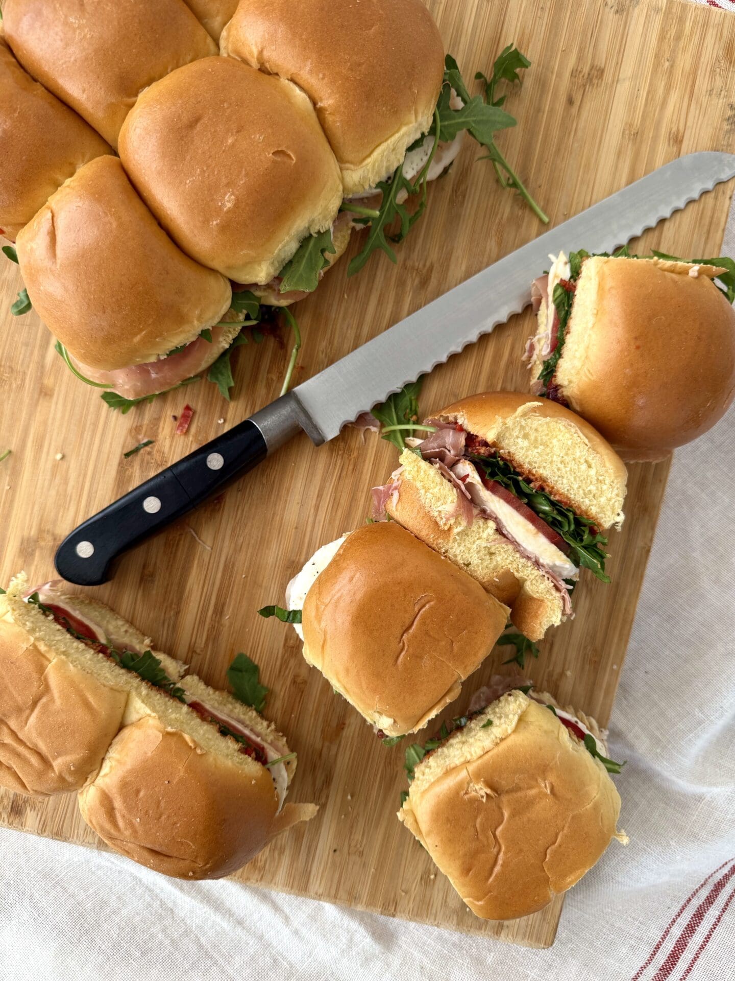 IMG_5048 Italian Antipasto Sliders are seen from above on a wood cutting board. A bread knife rests on the board and some of the sliders have been cut apart from the others. One slider is turned on its side, revealing its fillings.