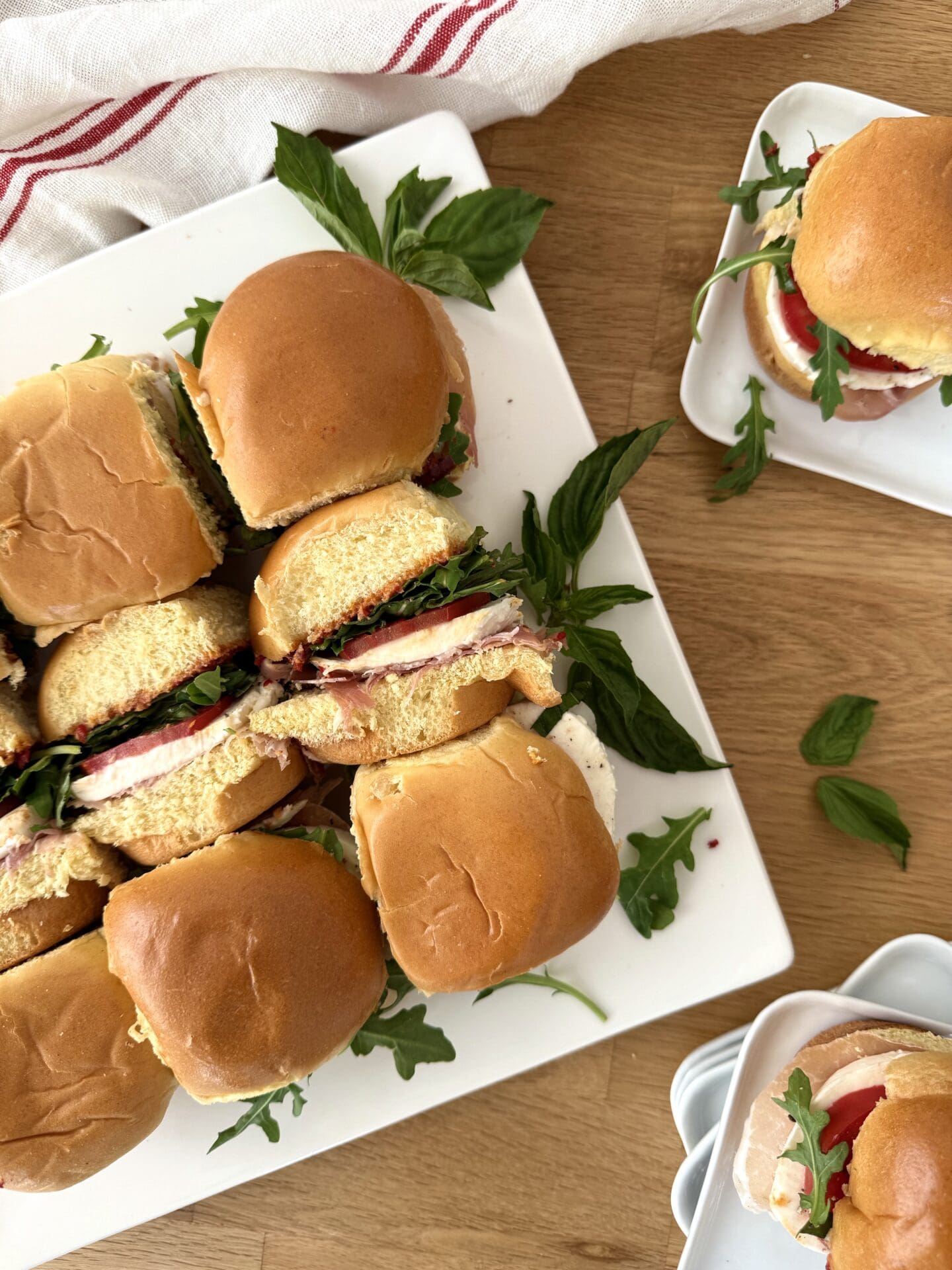 IMG_5055 A platter of Italian Antipasto Sliders is seen from above, surrounded by individual plates of sandwiches and fresh basil leaves.