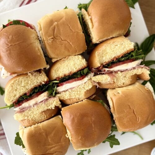 A platter of nine Italian Antipasto Sliders , arranged in 3 rows, is seen from above. The sliders in the centre row have been turned on their sides so you can see that they are stuffed with prosciutto, fresh mozzarella, tomatoes, basil and arugula.