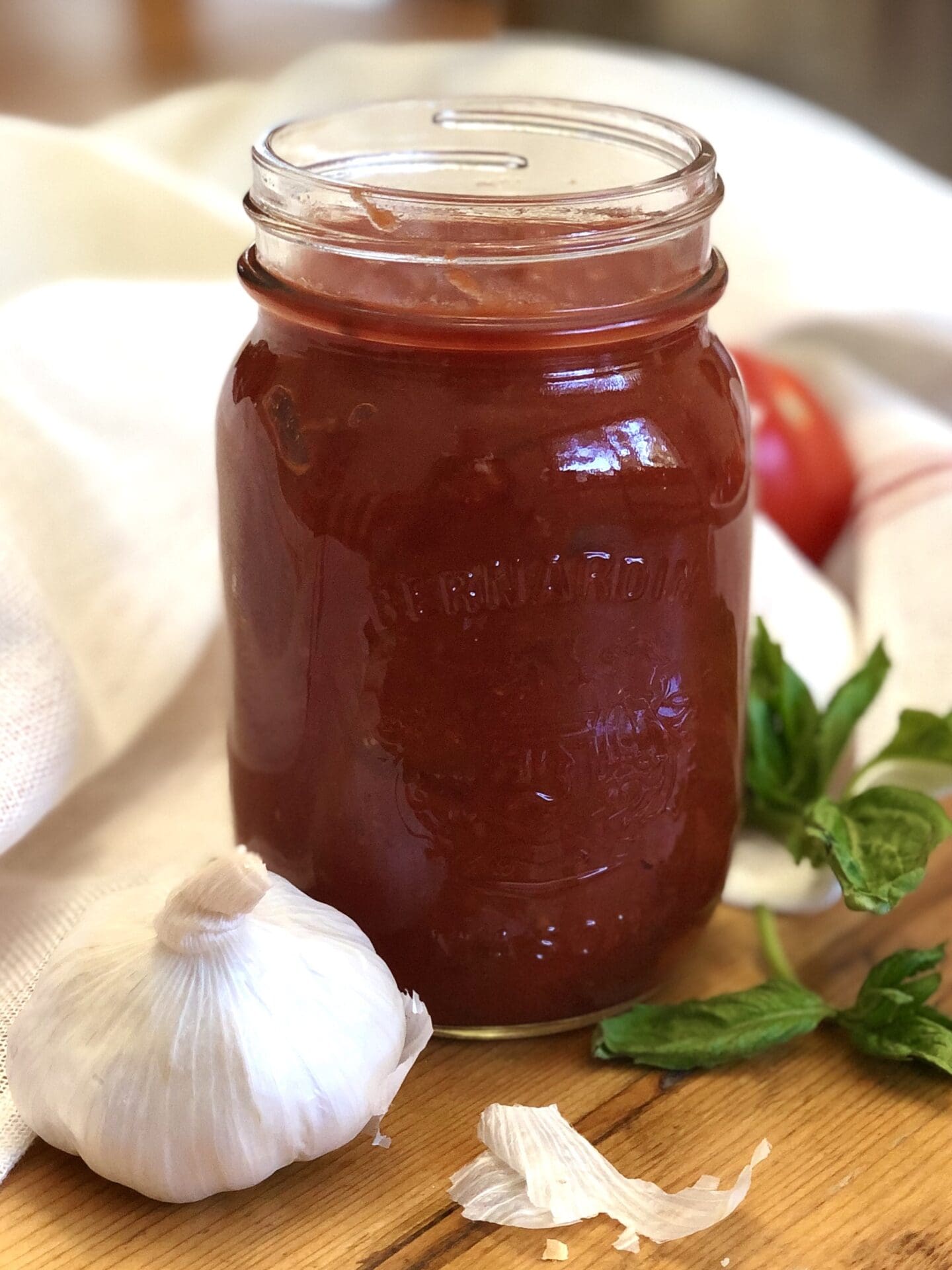 A jar of homemade Sarah's Marinara Sauce is seen on a aged wood table alongside fresh garlic, tomatoes and basil.