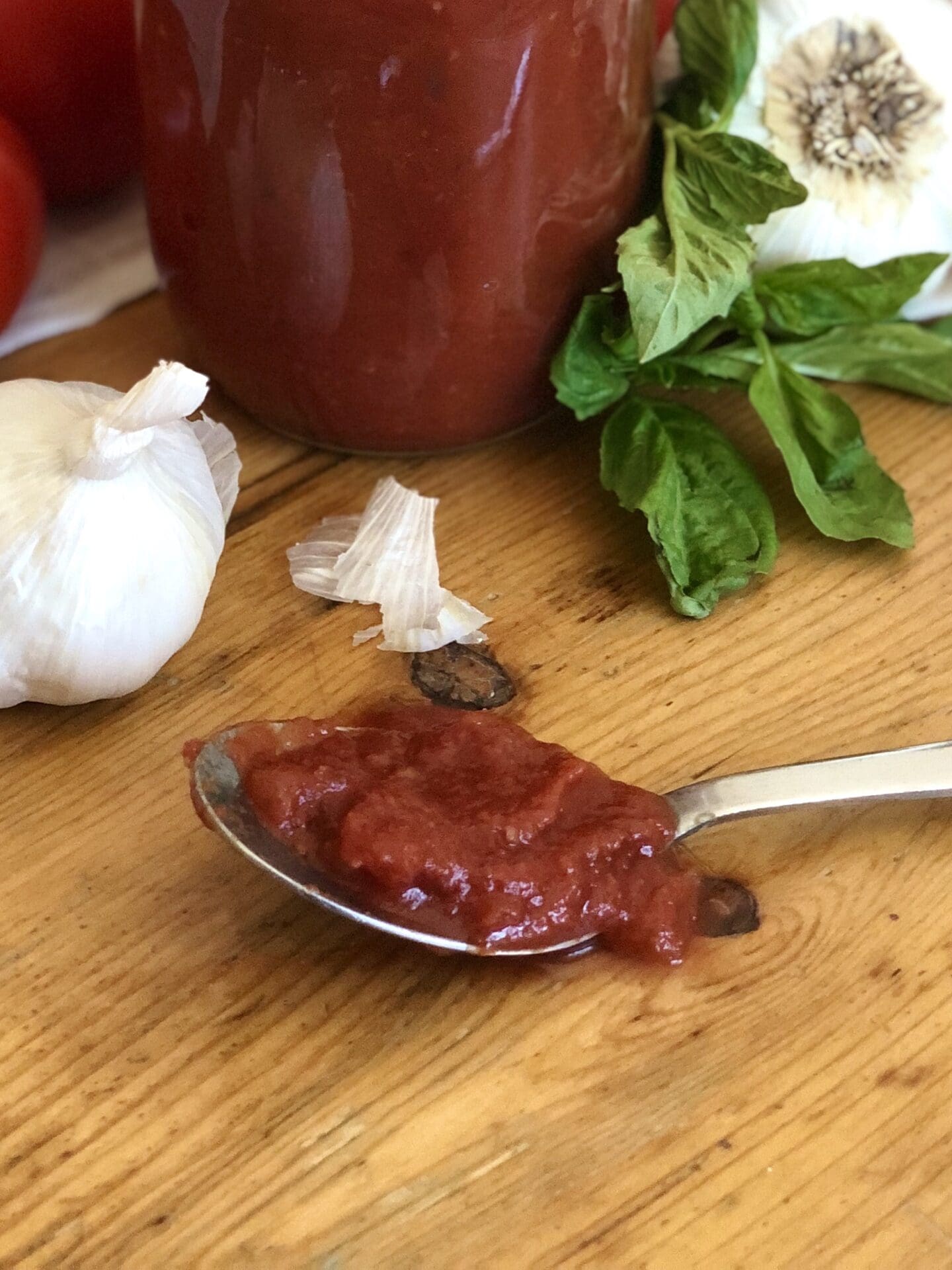 IMG_3158 A spoonful of thick and silky Sarah’s marinara sauce sits on a wooden table. A jar of the sauce, fresh springs of basil and whole heads of garlic are seen in the background.