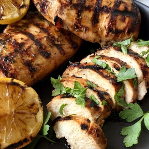 Sweet and Tangy Marinated Grilled Chicken is seen on a black platter with grilled lemon wedges and fresh parsley.