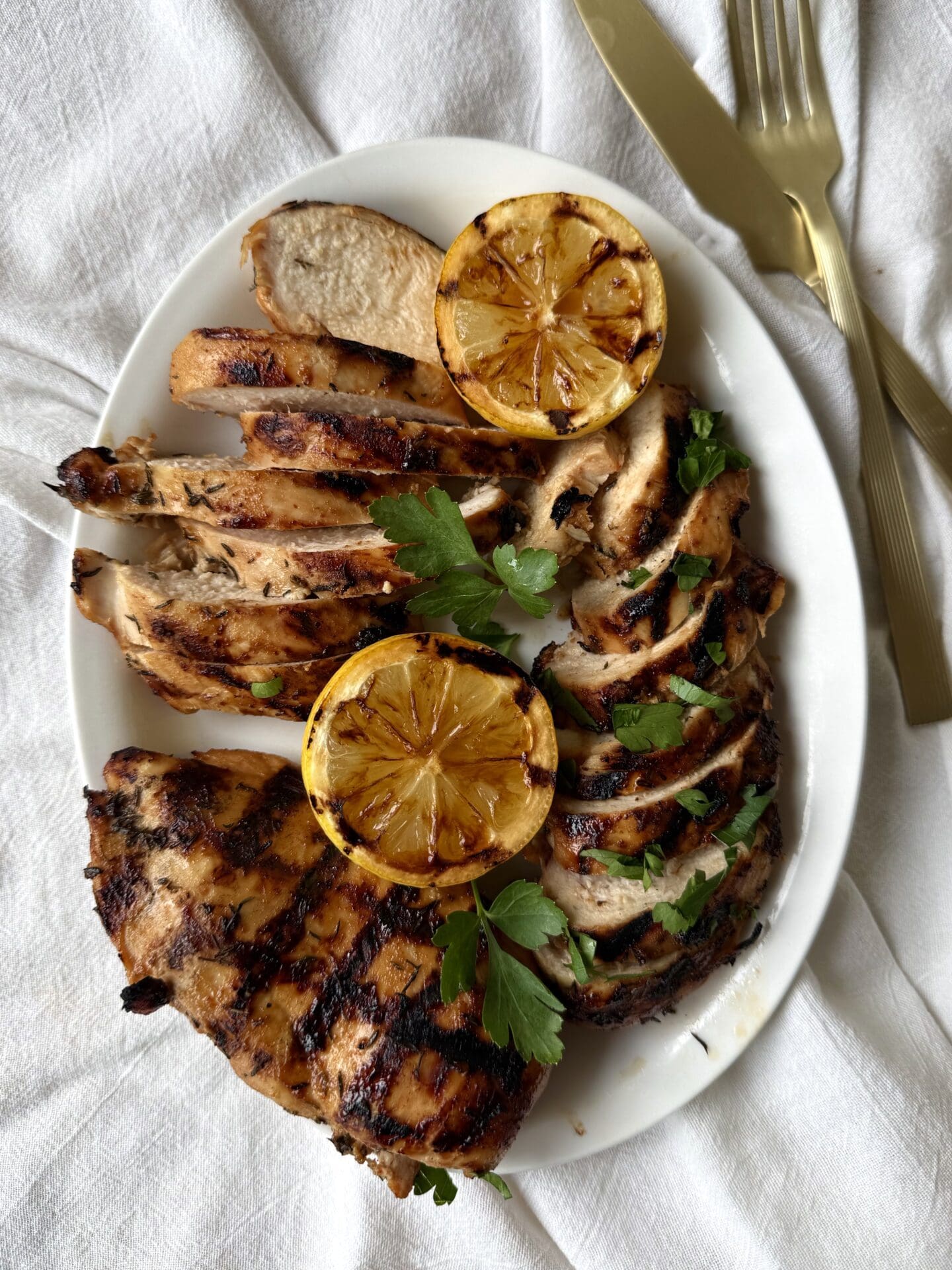 IMG_5172 A white platter is seen from above, filled with Sweet and Tangy Marinated Grilled Chicken. Some of the chicken breasts are sliced, whereas others are seen whole displaying their crosshatched grill marks. The platter rests on a white linen tablecloth. Gold coloured utensils are seen to the side.
