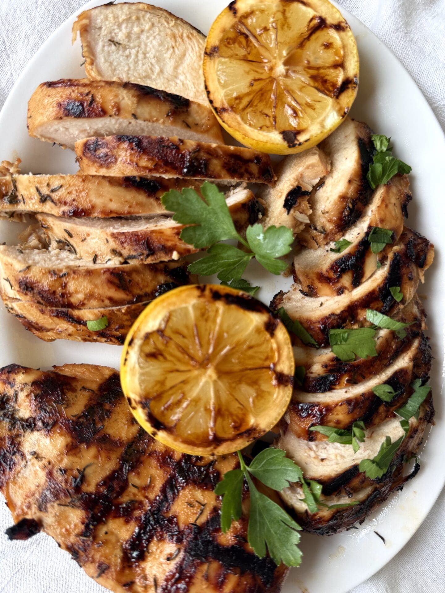 IMG_5175 A white platter of sweet and tangy marinated grilled chicken is seen from above, on a white linen tablecloth. The chicken is garnished with grilled lemon and fresh parsley.