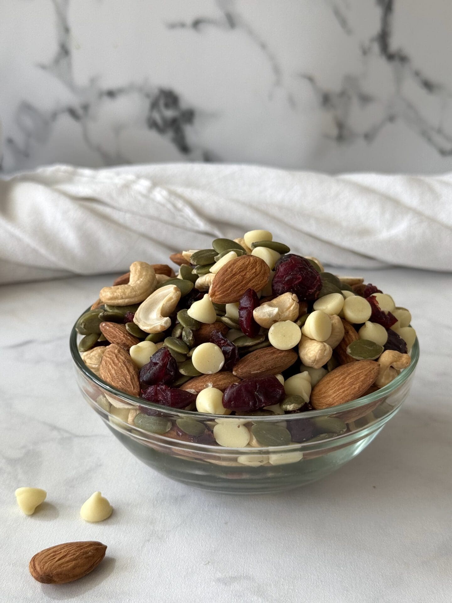 IMG_5213 A glass bowl of White Chocolate and Cranberry Trail Mix is seen on a white marble countertop. A few white chocolate chips and a whole almond sit beside the bowl.