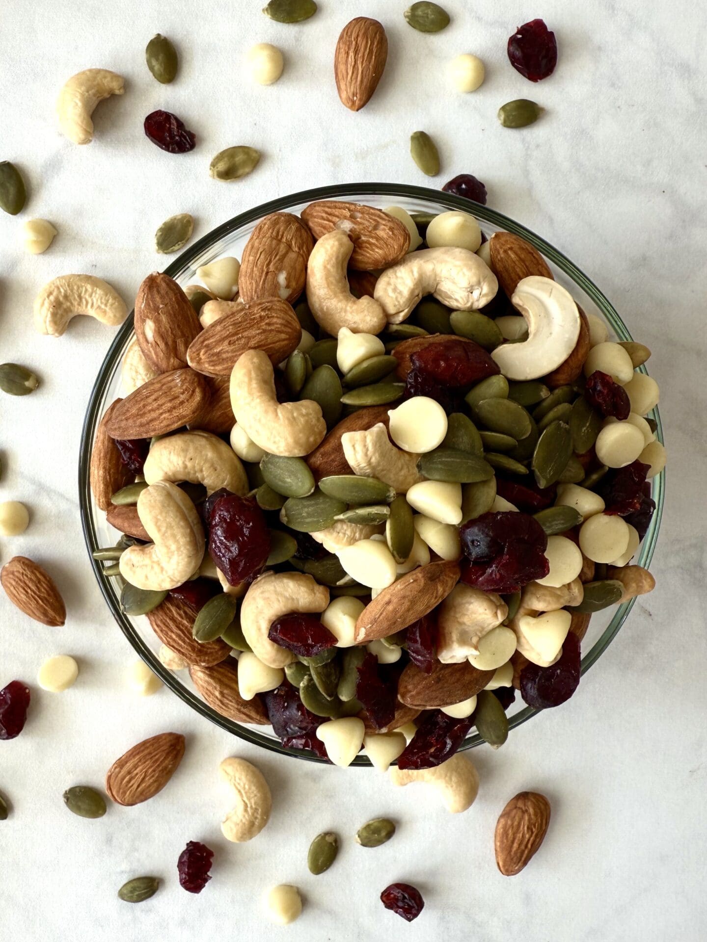 IMG_5220 A bowl of White Chocolate and Cranberry Trail Mix is seen from above. Scattered almonds, cashews, pumpkin seeds, dried cranberries and white chocolate chips can be seen scattered on the table around the bowl, on the left side.