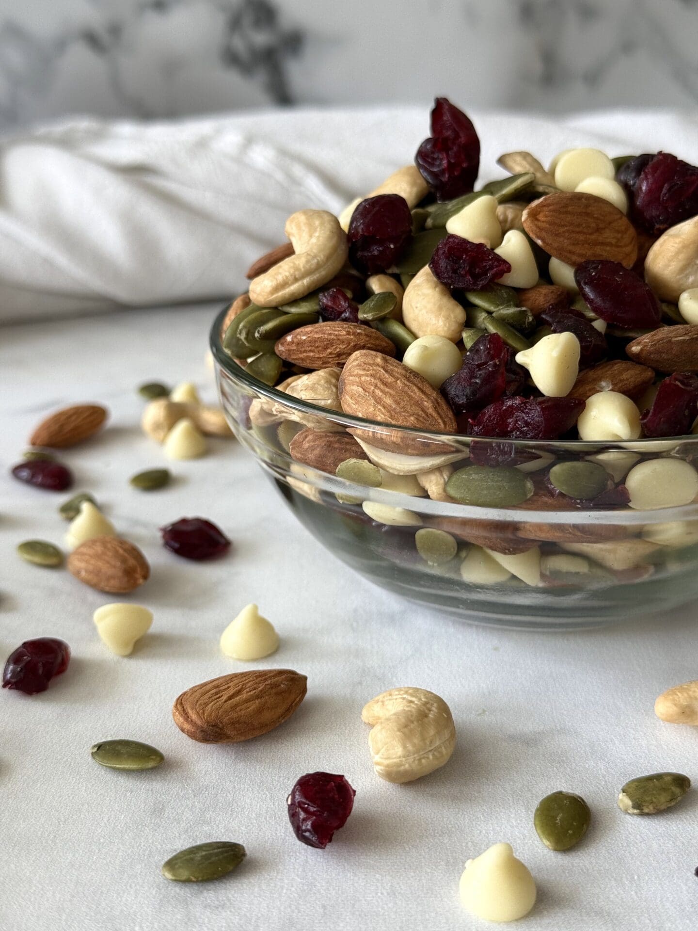IMG_5229 A glass bowl of White Chocolate and Cranberry Trail Mix is seen on a white marble countertop, surrounded by almonds, dried cranberries, cashews and pumpkin seeds.