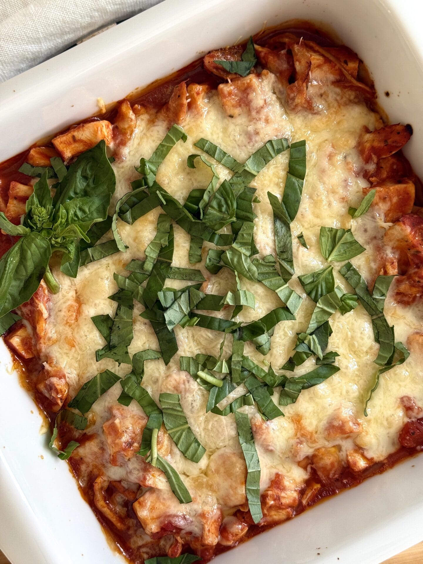 A white baking dish of Healthier Chicken Parmesan is seen from above, covered in melted cheese and sprinkled with fresh basil.