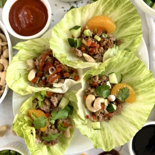 A plate of Ground Turkey Lettuce Wraps is seen from above, surrounded by dishes of chopped cucumber, spring onions, hoisin sauce, cashews and sriracha sauce.
