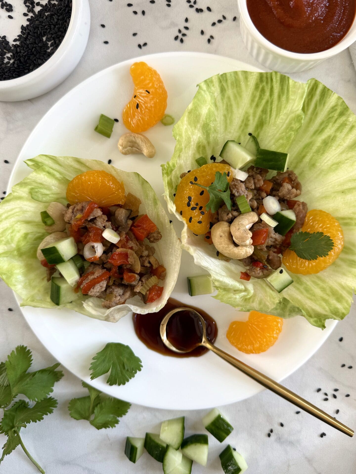A dinner plate is seen from above with two turkey lettuce wraps. The lettuce wraps are garnished with mandarin orange slices, chopped cucumber, spring onions, sesame seeds and hoisin sauce.