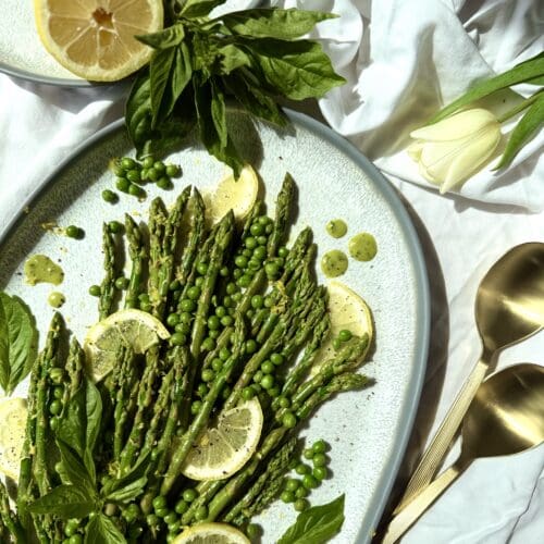 A platter of Lemon Basil Asparagus and Peas is seen from above, lit with bright spring sunshine. Lemons and fresh basil surround the platter that rests on a white table cloth. Pale yellow tulips and gold serving spoons complete the image.