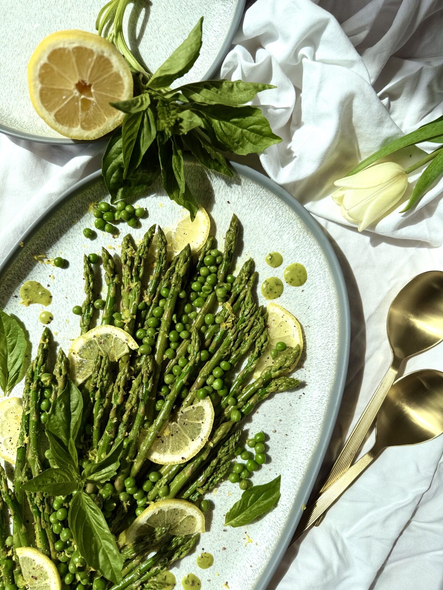 IMG_5388 A platter of Lemon Basil Asparagus and Peas is seen from above, lit with bright spring sunshine. Lemons and fresh basil surround the platter that rests on a white table cloth. Pale yellow tulips and gold serving spoons complete the image.