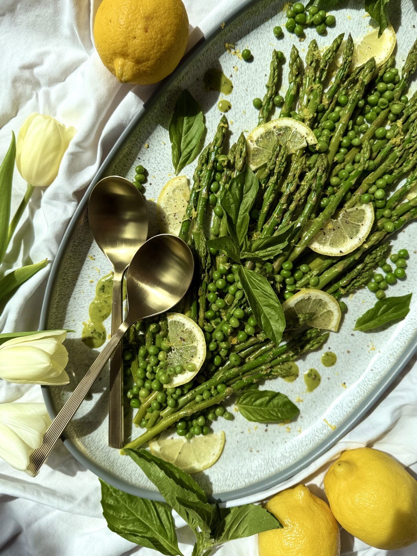 IMG_5404 A platter of Lemon Basil Asparagus and Peas is seen from above on a white table cloth. The vegetables are garnished with lemon wedges, fresh basil leaves, vinaigrette drips and freshly ground black pepper. A set of gold serving spoons rest on the platter. Tulips and lemons complete the image.