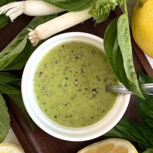 A bowl of Homemade Lemon Basil Dressing sits on a dark wood tray, surrounded by lemons, fresh basil leaves and green onions.