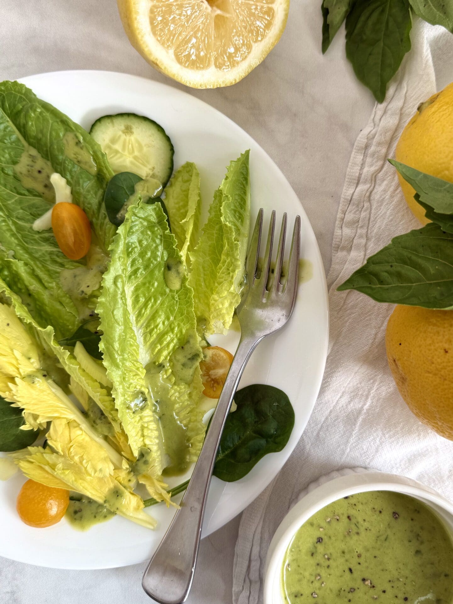 IMG_5421 A fresh green salad is seen on a white background, with a bowl of Lemon Basil Vinaigrette served on the side. Fresh lemons and basil leaves complete the photo.