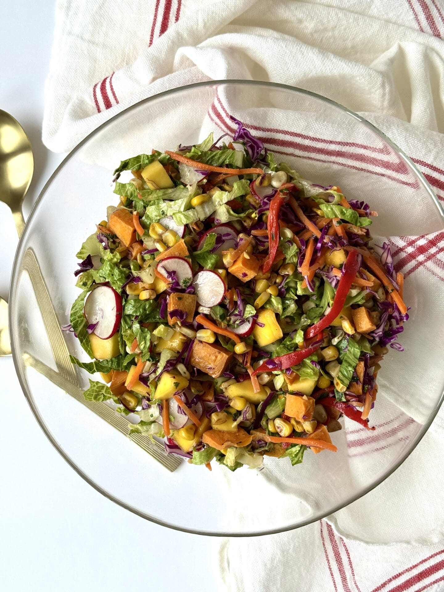 A glass bowl filled with Mexican Chopped Salad is seen from above on a white background.  A red and white striped cloth and gold coloured salad server surround the bowl.