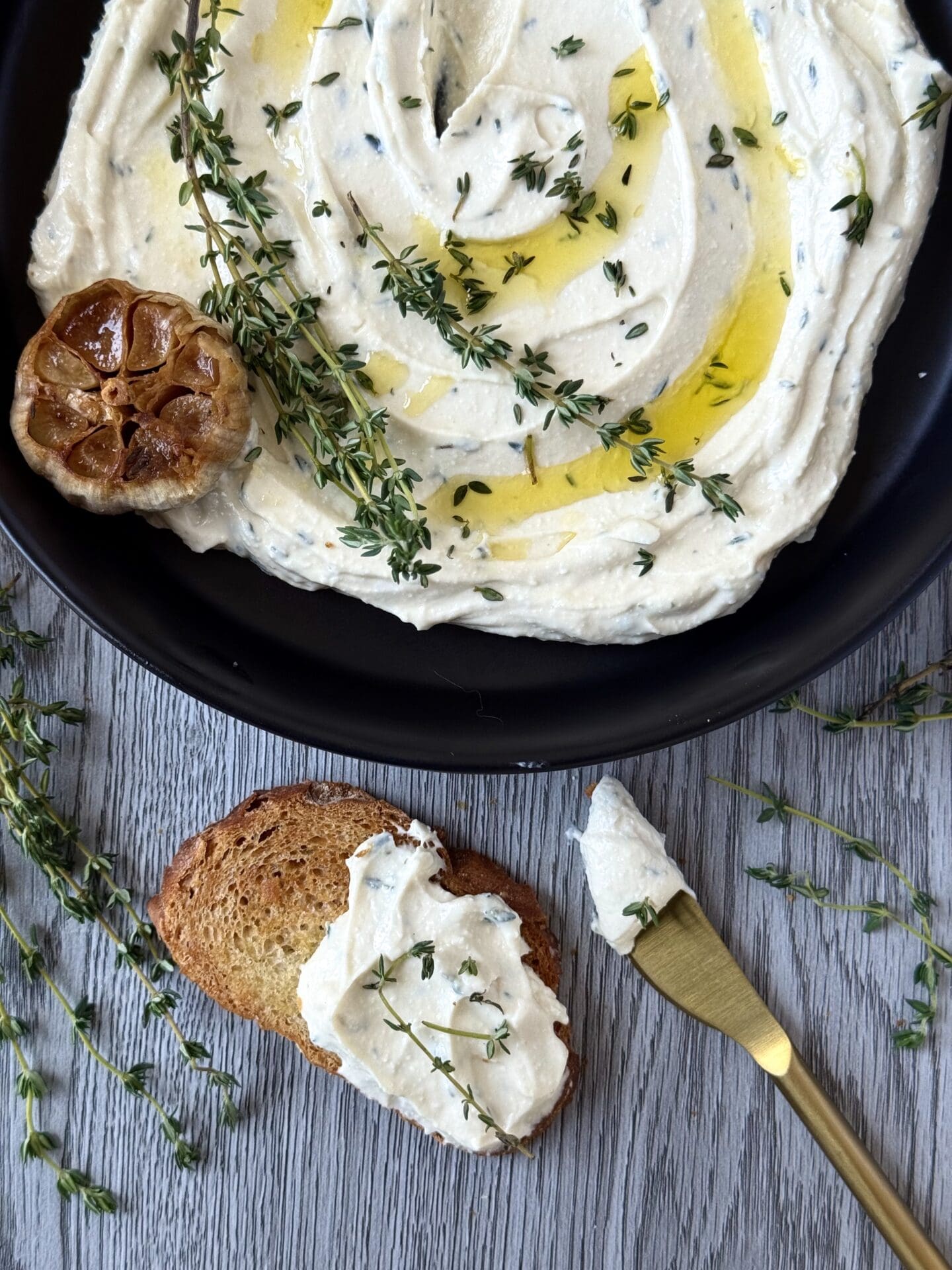 A black dish is seen from above, filled with swirled Whipped Feta with Roasted Garlic and Thyme.  In front of the bowl sits a slice of toasted baguette slathered in the dip.  A gold spreader, a head of roasted garlic and sprigs of fresh thyme complete the picture.