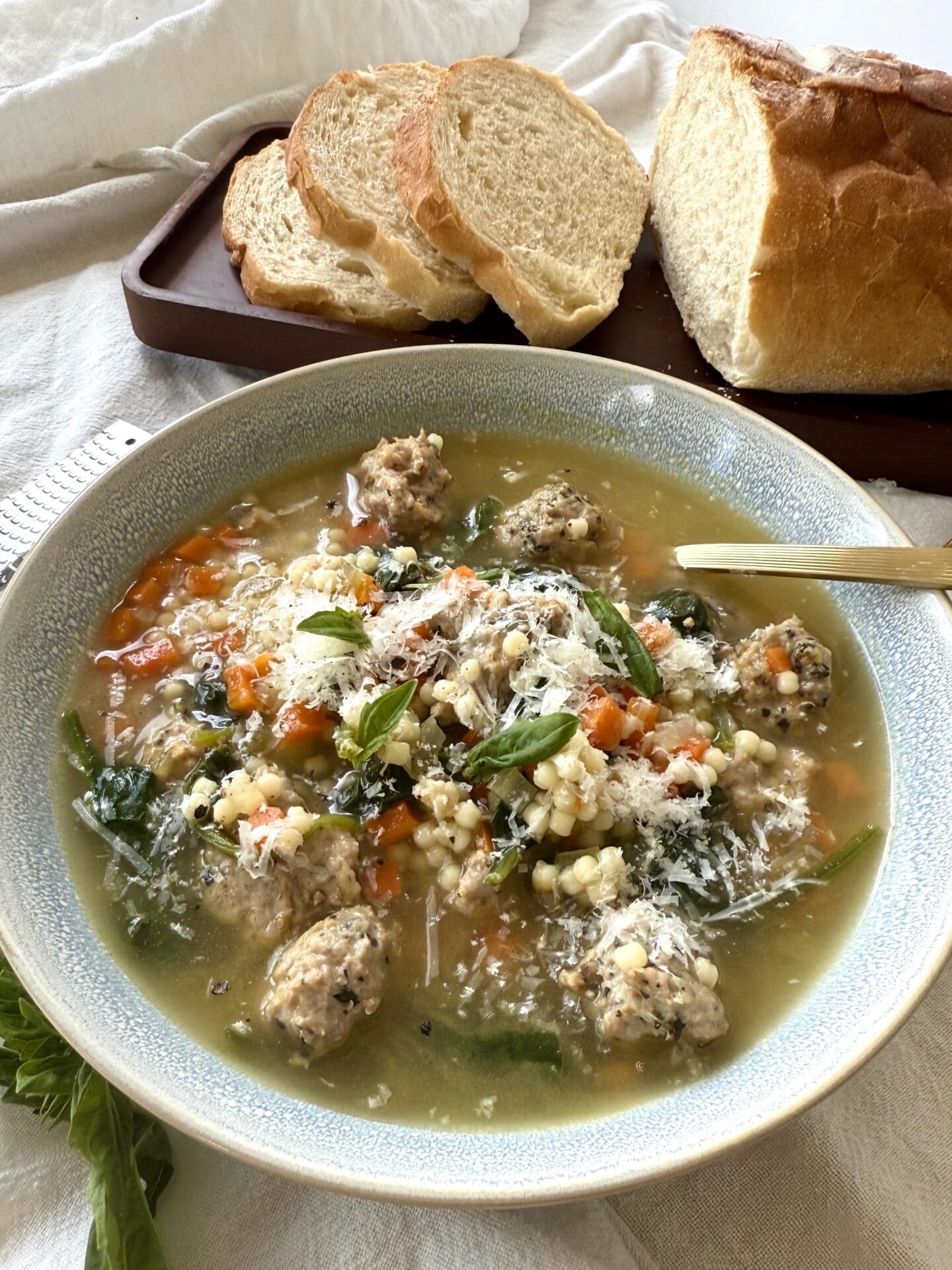 A bowl of Healthier Italian Wedding Soup sits on a table, laden with roasted turkey meatballs and garnished with freshly grated pecorino Romano cheese and basil leaves.  A loaf of fresh Italian bread sits in the background.