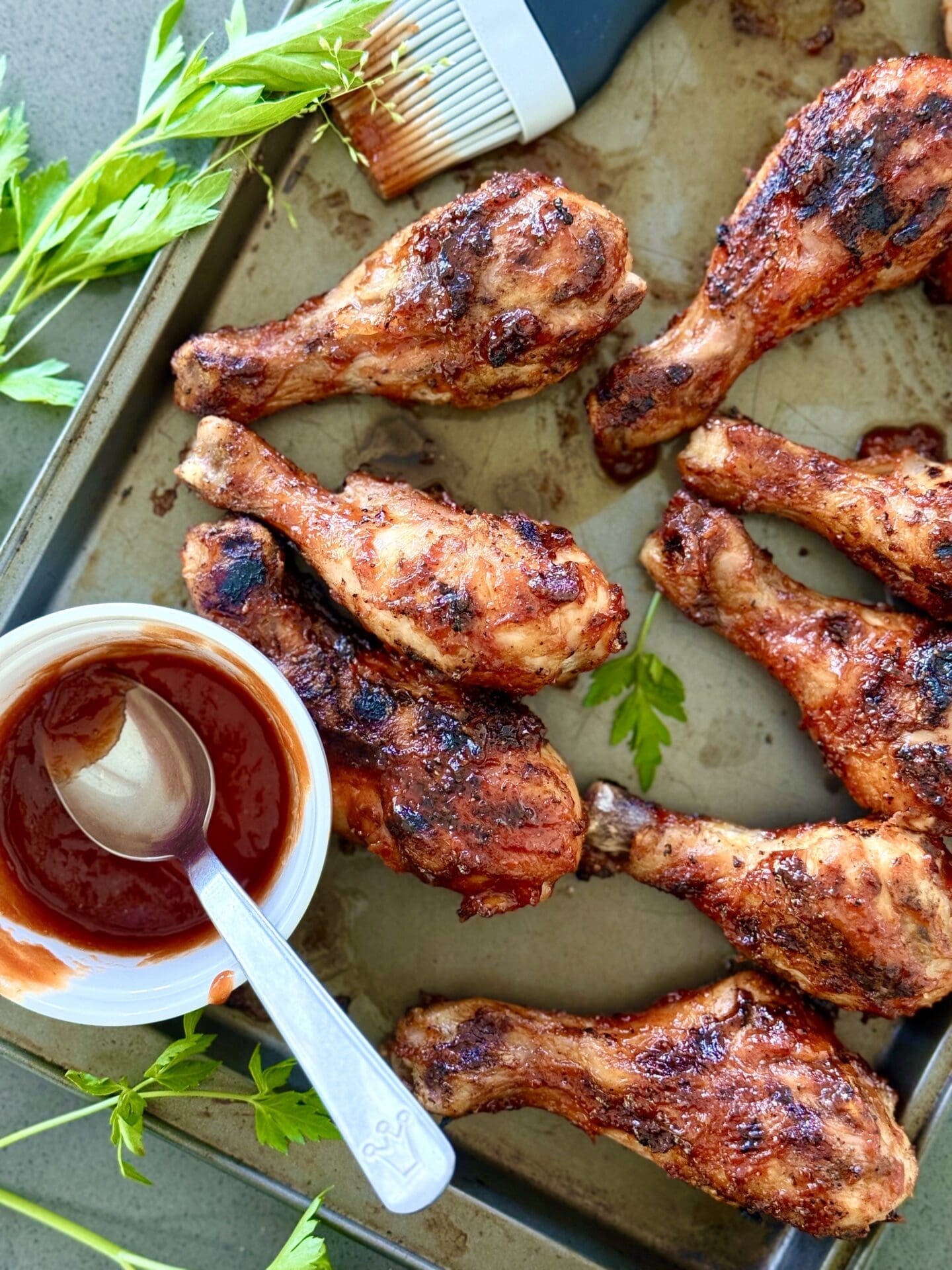 IMG_5943 A baking sheet is filled with Smokey Sweet Grilled Chicken Legs. A bowl of homemade barbecue sauce and a basting brush are seen on the edges.