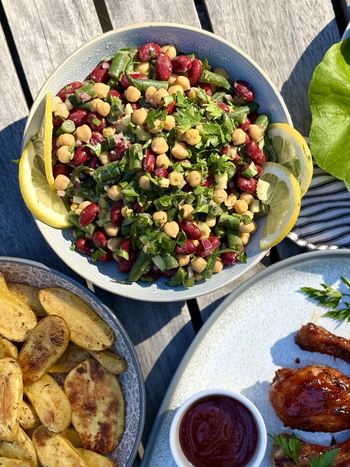 IMG_5949 A bowl of Marinated Mediterranean Bean Salad is seen from above on an outdoor table, as part of a backyard barbecue dinner. The salad is surrounded by platters of grilled chicken and roasted potatoes.