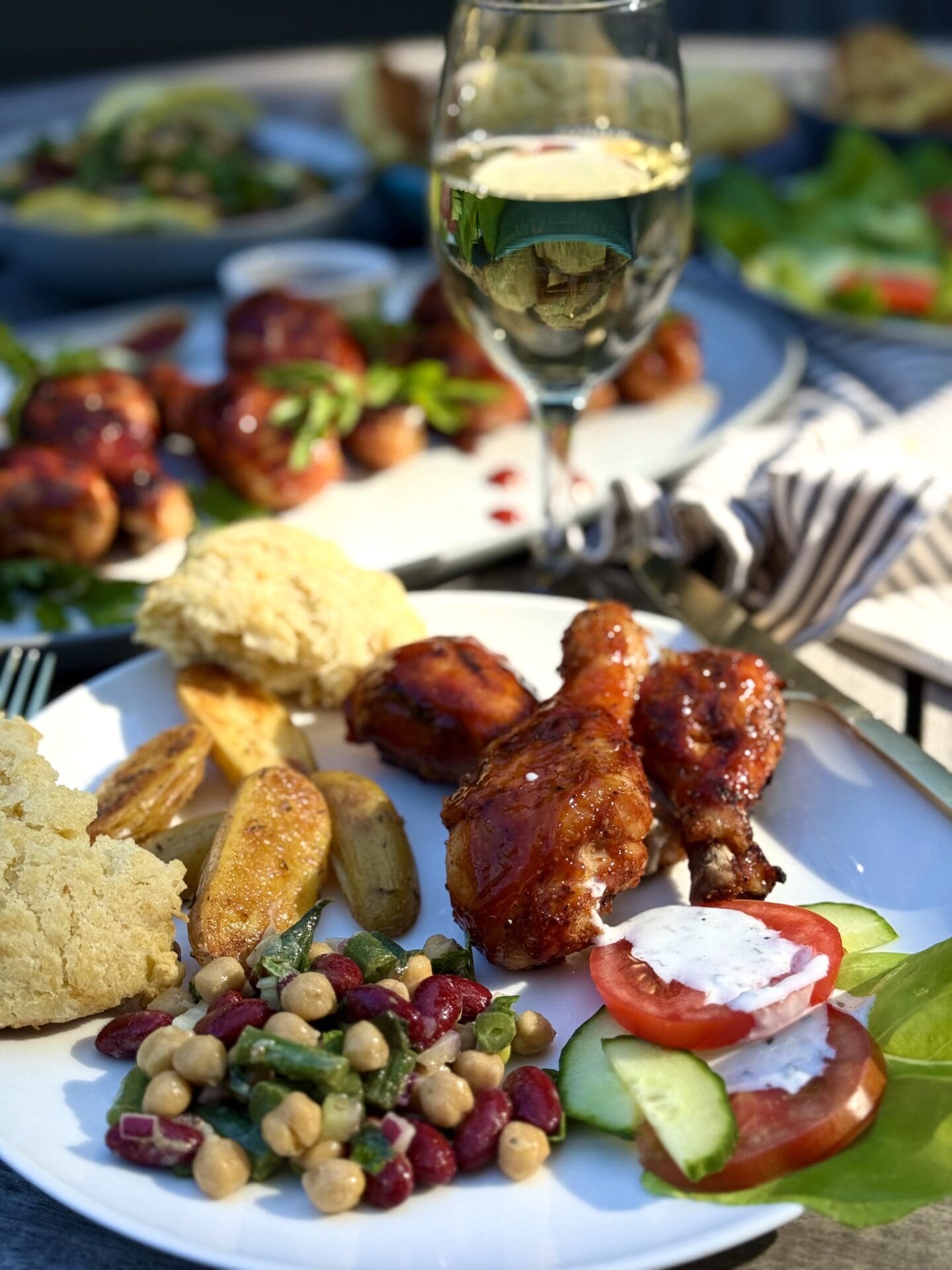 IMG_5961 A dinner plate of Marinated Mediterranean Bean Salad with roasted potatoes, salad and grilled chicken is seen on a picnic table. A glass of cold white wine and platters of food are seen in the background.