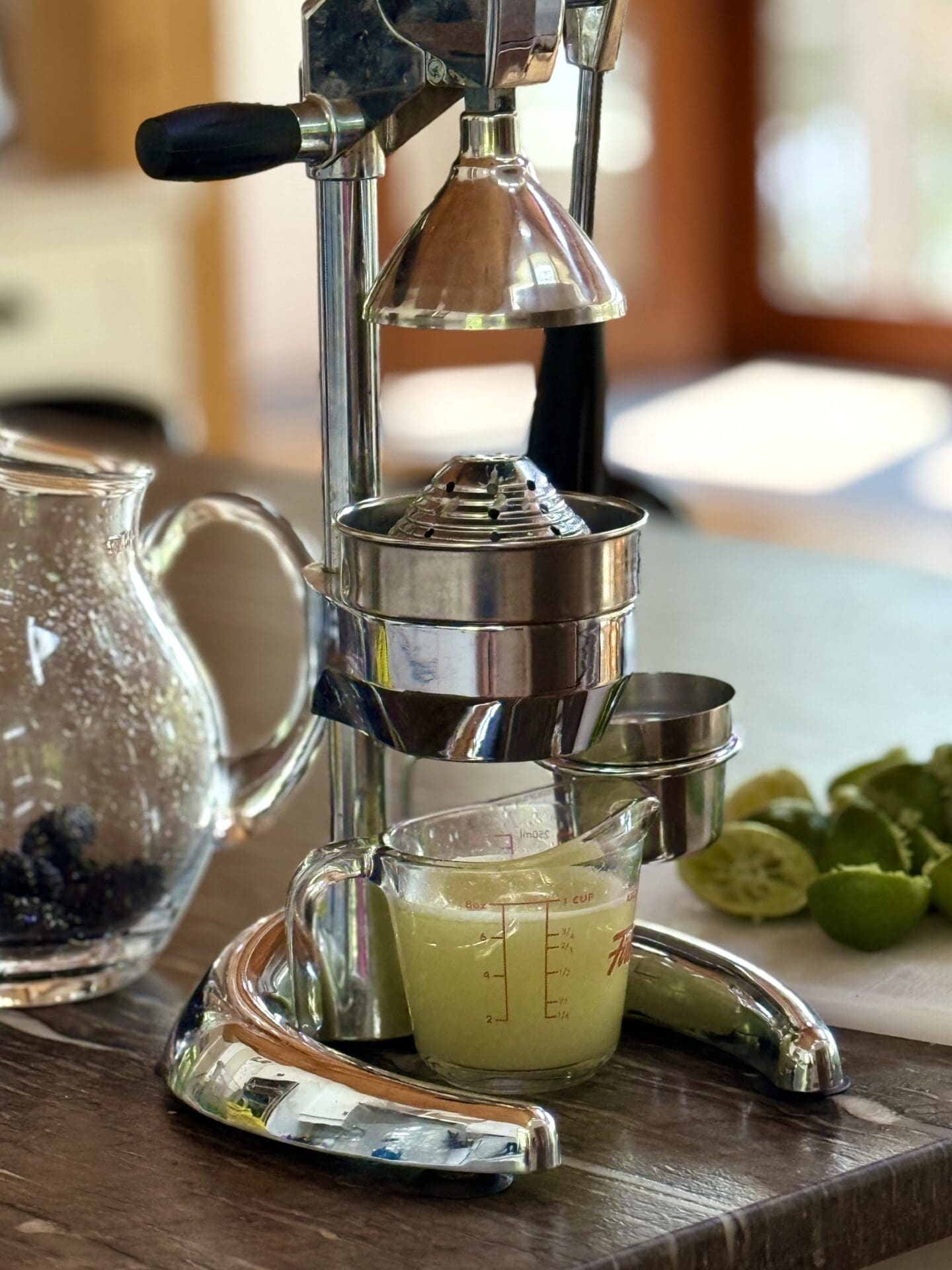 IMG_6456 A citrus press sits on a grey stone countertop where it has been pressing fresh limes. A pitcher full of lime juice sits under the press and sunshine streams in through a screen door in the background.