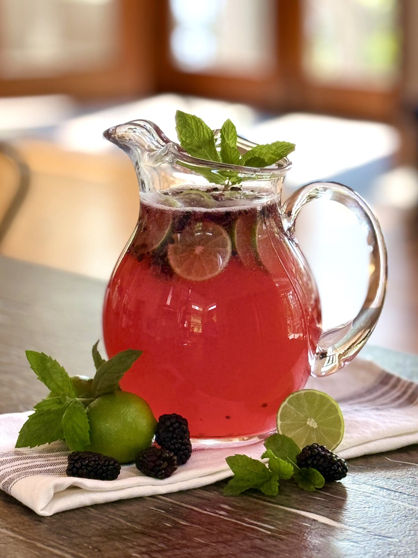 IMG_6464 A glass pitcher full of Blackberry Mojito Mocktail sits on a table, garnished with blackberries, lime wedges and fresh mint. Sunshine streams in through the screen doors in the background.