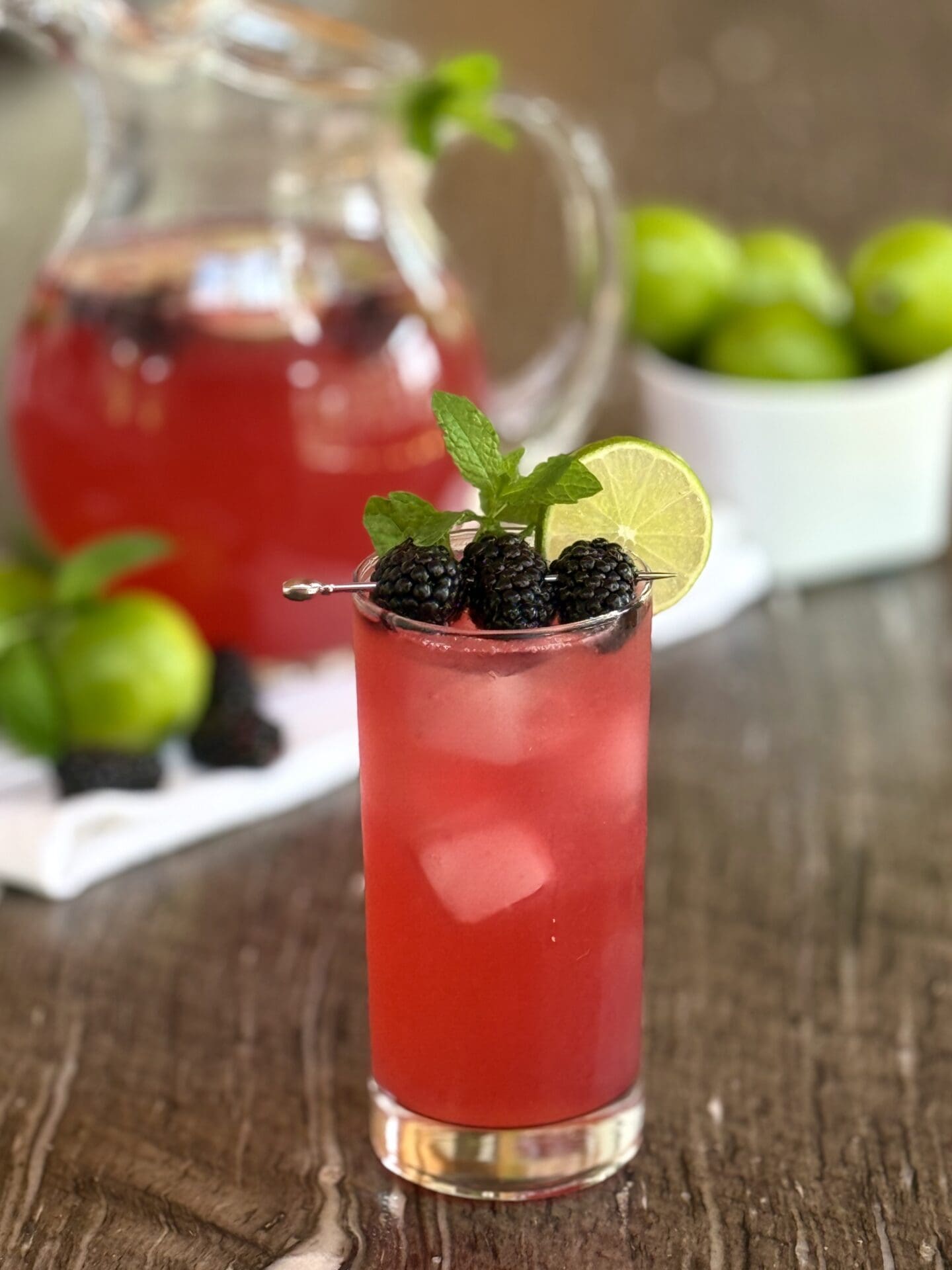 Blackberry Mojito Mocktail is served over ice in a tall glass. In the background a pitcher of the vibrant alcohol-free drink is surrounded by limes, juicy blackberries and sprigs of fresh mint.