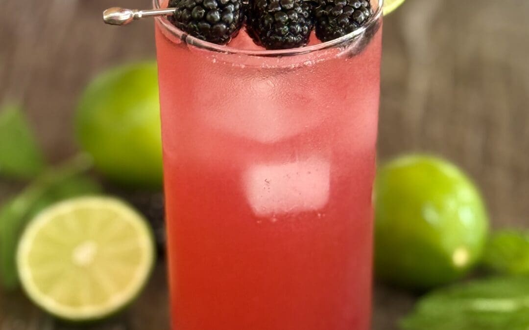 A tall glass of Blackberry Mojito Mocktail sits on a grey stone countertop , garnished with plump blackberries, lime wheels and fresh mint sprigs.