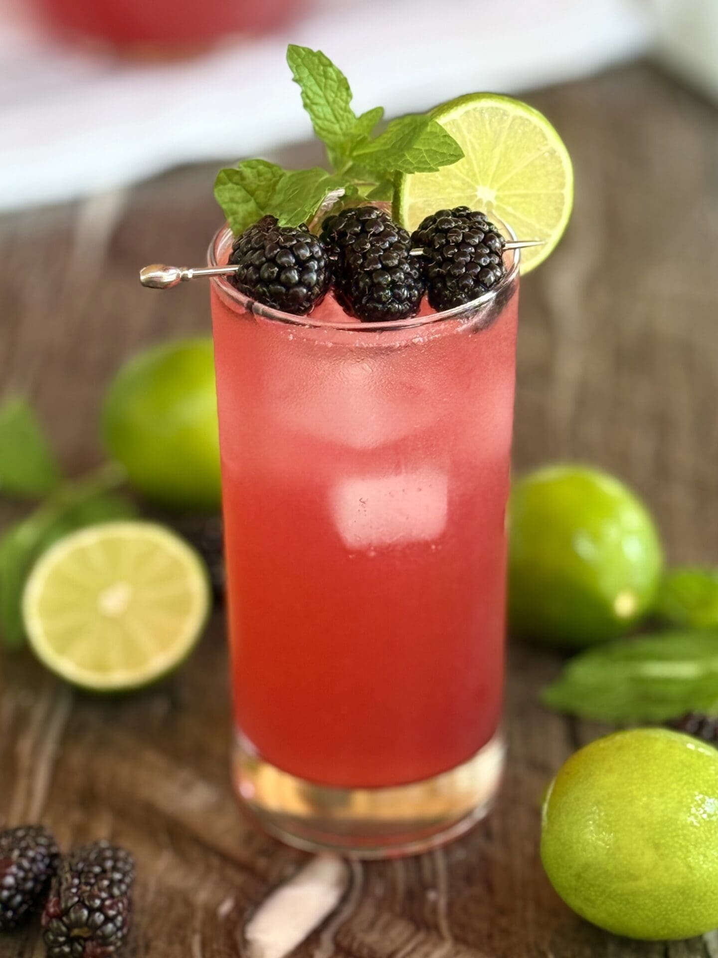 A tall glass of Blackberry Mojito Mocktail sits on a grey stone countertop , garnished with plump blackberries, lime wheels and fresh mint sprigs.