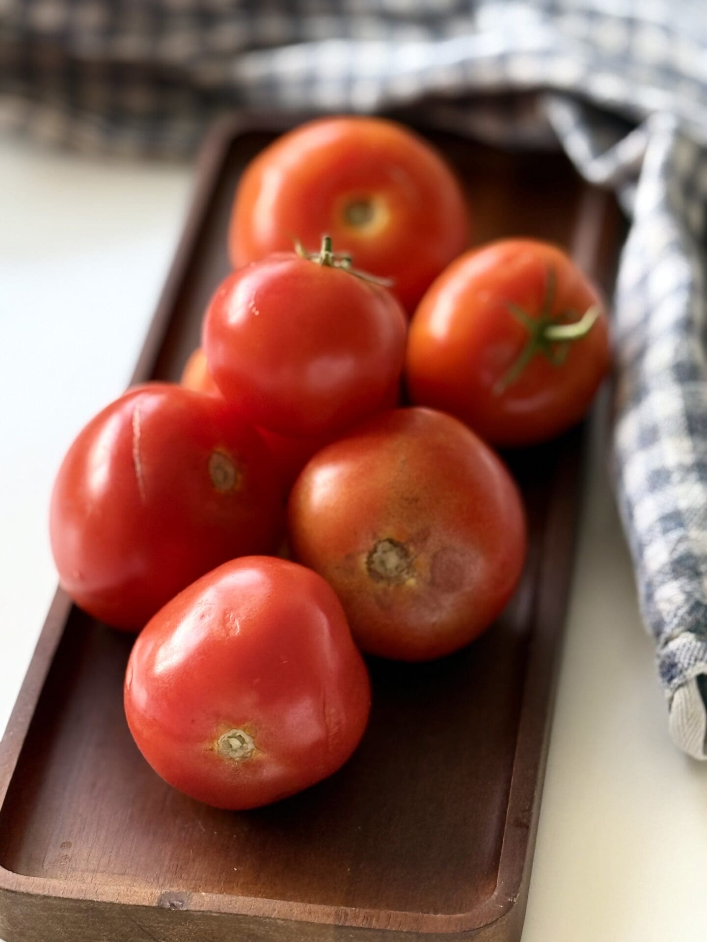 IMG_6804 A wood tray is stacked high with garden fresh tomatoes of varying sizes. A checkered cloth is seen in the background.