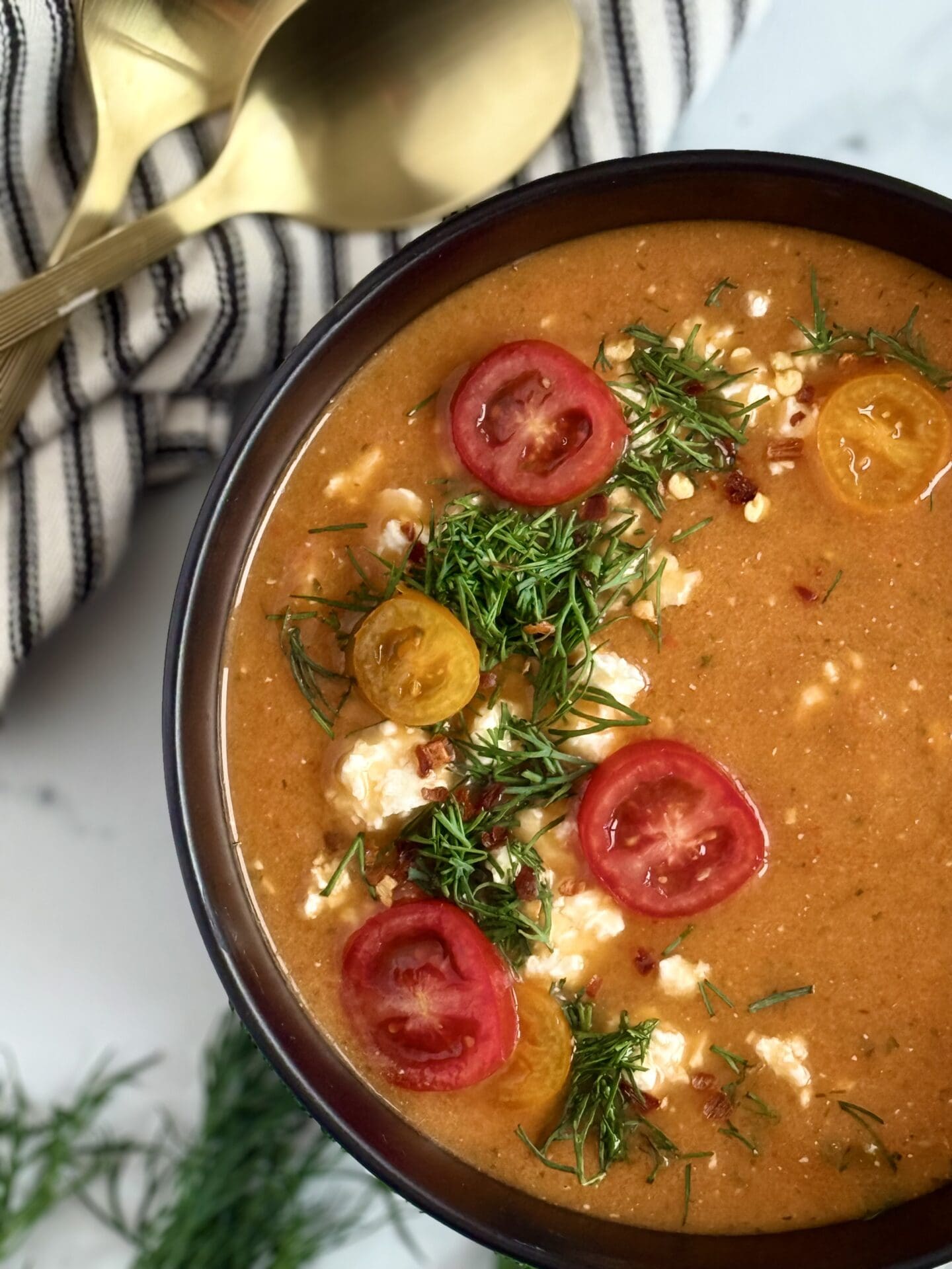 A black bowl filled with roasted tomato and feta soup is seen from above, beautifully garnished with fresh dill, crumbled feta cheese and sliced cherry tomatoes.