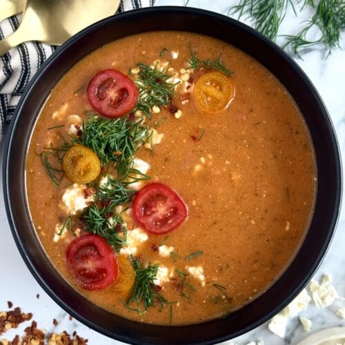 A black bowl filled with roasted tomato and feta soup is seen from above. The soup is garnished with a semi-circle of fresh chopped dill, crumbled feta cheese and sliced cherry tomatoes. Fresh dill, feta, chili flakes and gold spoons surround the bowl.
