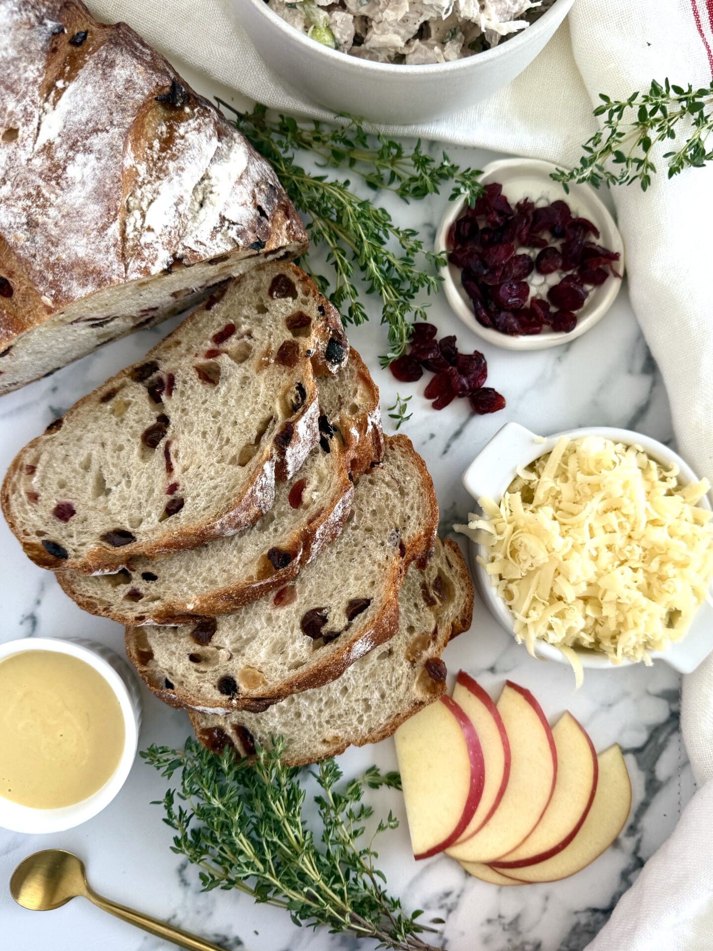 IMG_6869 The ingredients for Chicken and Apple Melt Sandwiches with Smoked Cheddar are seen from above, resulting on a white marble countertop. A loaf and slices of cranberry-studded bread are surrounded by apple slices, maple mustard, grated smoked cheddar and fresh thyme.