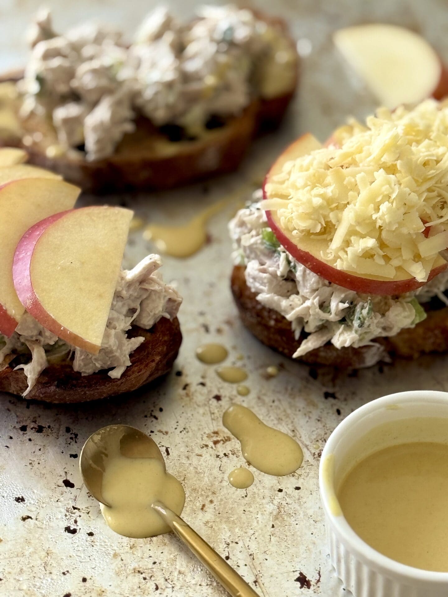 Chicken and Apple Melt Sandwiches are seen being prepared before going in the oven. Apple slices and drips of maple mustard surround the sandwiches while they rest on a metal baking sheet.
