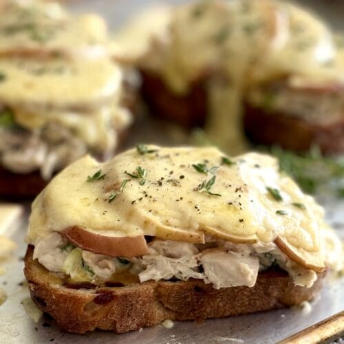 Chicken and Apple Melts with Smoked Cheddar are seen hot from the oven with melting cheese dripping down over the edges of the sandwiches.